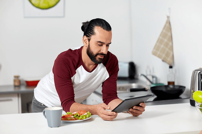 Two people in a bright kitchen slicing cucumber, surrounded by fresh fruits and vegetables on the counter