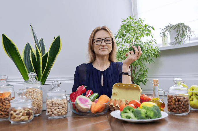 Woman holding avocado at table with vegetables, nuts, grains, eggs, oils, and plants promoting healthy eating