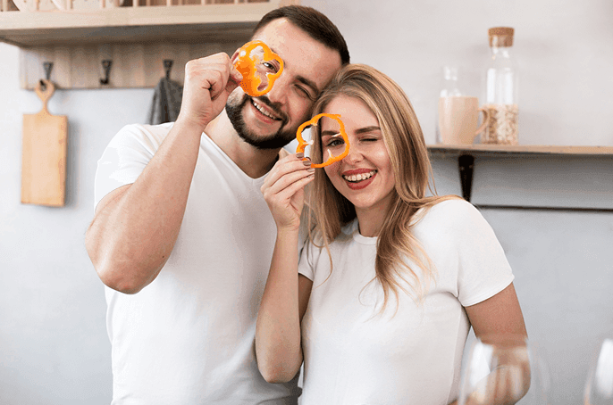 Two people in a kitchen playfully holding orange bell pepper slices over their eyes, smiling together