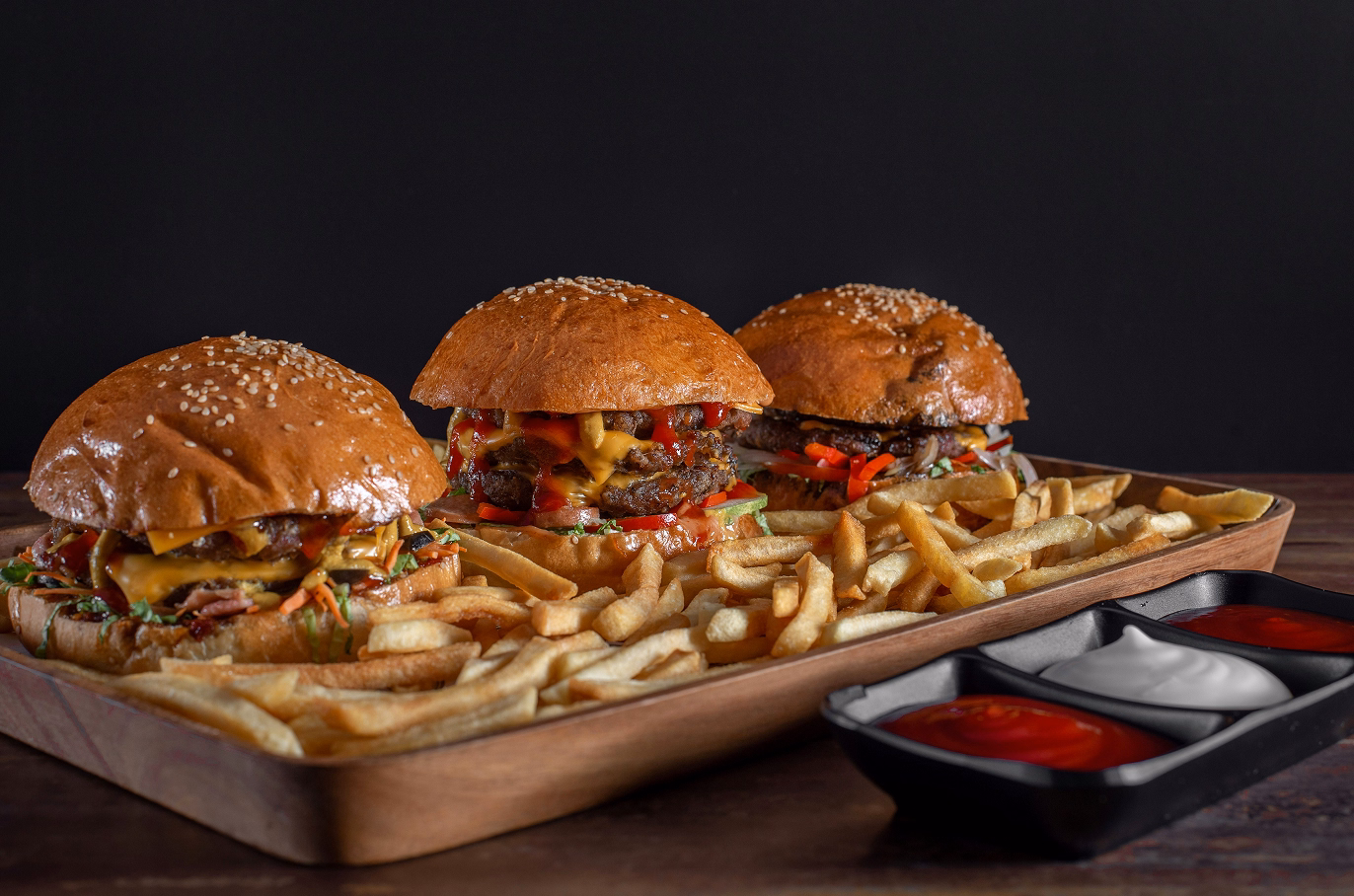 Wooden tray with three cheeseburgers, fries, and condiments against a black background