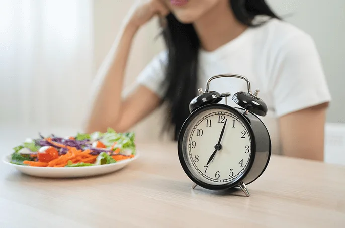 Black alarm clock at 7:03 beside person eating fresh salad with tomatoes, carrots, and cabbage